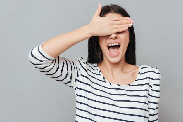 Mulher jovem feliz em pé sobre a superfície cinza e cobrindo os olhos.