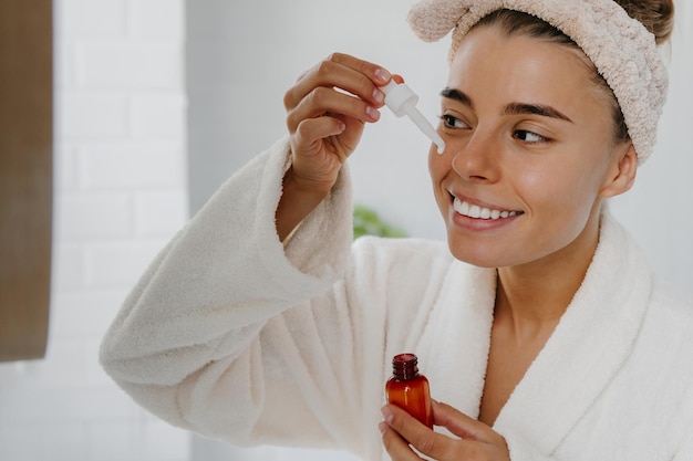 Mulher jovem feliz em roupão de banho aplicando soro cosmético no rosto enquanto está de pé no banheiro
