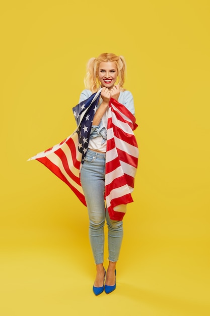 Mulher jovem feliz em roupas jeans, segurando a bandeira dos eua sobre fundo amarelo.