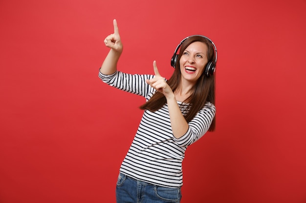 Mulher jovem feliz em roupas listradas com fones de ouvido sem fio, ouvindo música, apontando o dedo indicador de lado, isolado no fundo vermelho. emoções sinceras de pessoas, conceito de estilo de vida. simule o espaço da cópia.