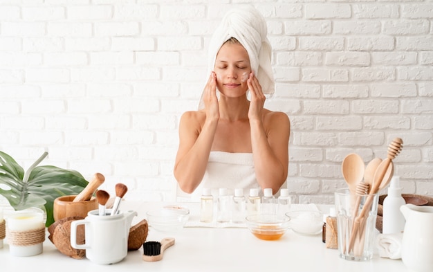 Mulher jovem feliz em toalhas de banho brancas, aplicar o creme na pele do rosto, fazendo precedências de spa