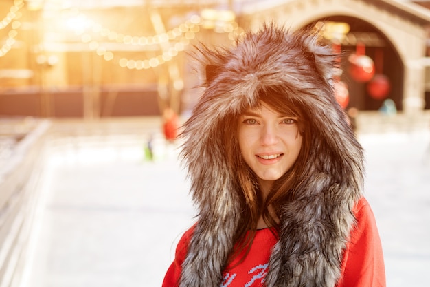 Mulher jovem feliz em um chapéu de lobo no inverno na pista de gelo posa com um suéter vermelho do lado de fora à tarde