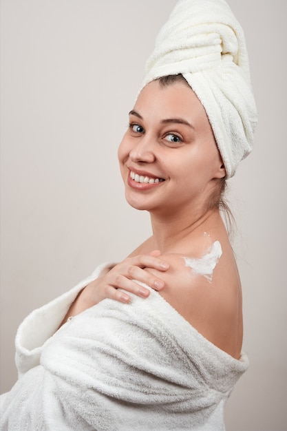 Mulher jovem feliz em um manto branco, com uma toalha na cabeça, passando creme no ombro