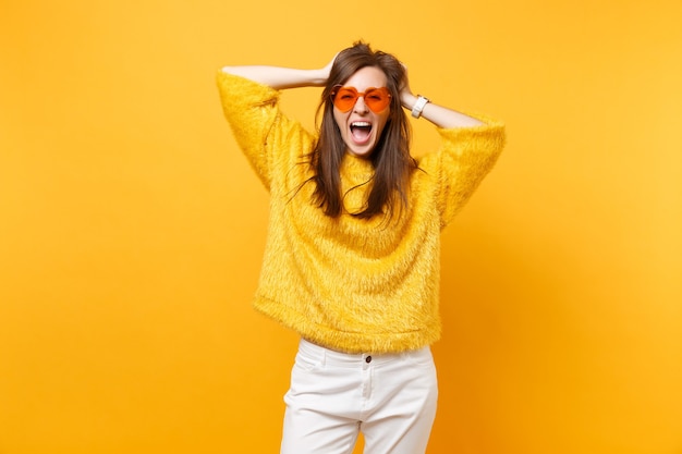 Mulher jovem feliz em um suéter de pele, calça branca, óculos coração laranja agarrados à cabeça, gritando isolado em fundo amarelo brilhante. emoções sinceras de pessoas, conceito de estilo de vida. área de publicidade.