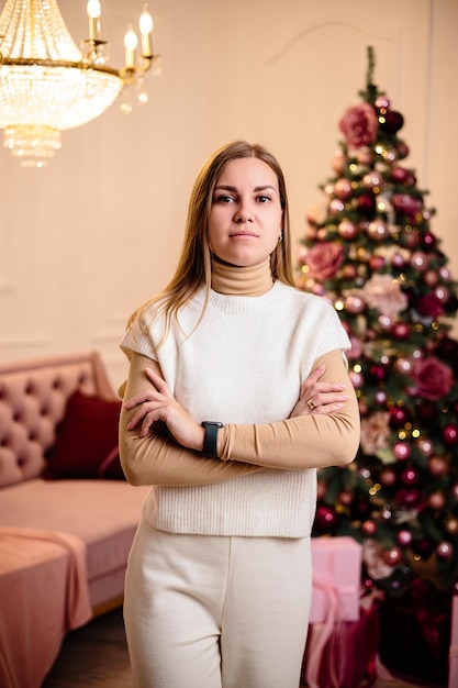 Mulher jovem feliz em um terno branco de malha no fundo da atmosfera festiva de ano novo de árvores de Natal