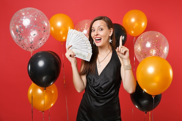 Mulher jovem feliz em um vestido preto, apontando o dedo indicador para cima segurando um pacote de muitos dólares, dinheiro em balões de ar de fundo vermelho. dia da mulher, feliz ano novo, conceito de festa de feriado de maquete de aniversário.