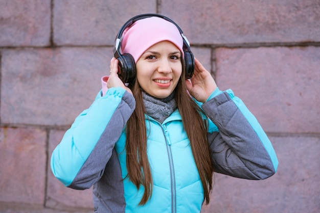 Mulher jovem feliz em uma jaqueta quente e chapéu rosa, ouvindo música em fones de ouvido contra o background.