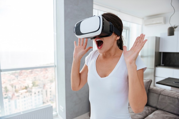 Mulher jovem feliz espantada com as mãos levantadas e a boca aberta usando googles de realidade virtual em casa