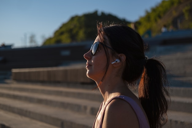 Mulher jovem feliz fitness em óculos de sol terminar o exercício ao ar livre na cidade. descansar. Desfrute de ouvir música em fones de ouvido. pôr do sol. estilo de vida saudável. liberdade. luz de contorno.