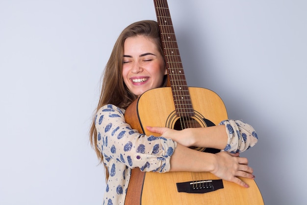 Mulher jovem feliz na blusa bege abraçando uma guitarra em fundo cinza no estúdio
