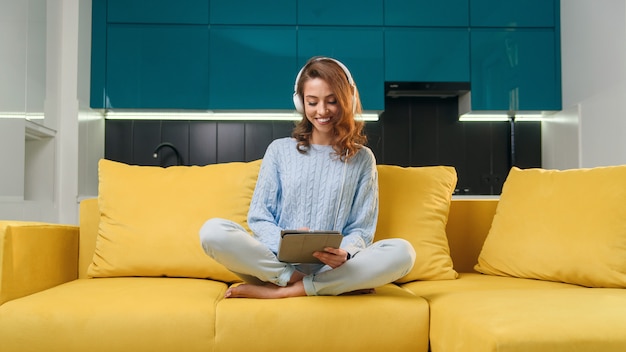 Mulher jovem feliz na camisola casual azul, sentado no sofá confortável e usa o tablet pc para ouvir música na cozinha contemporânea.