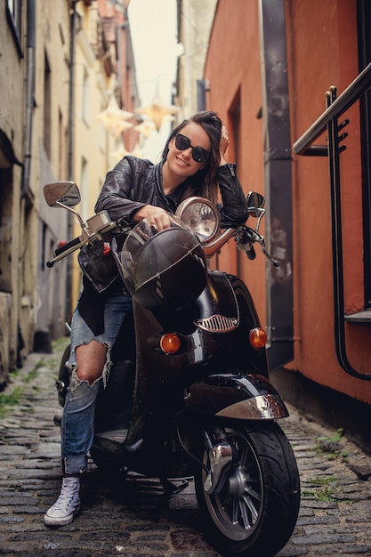 Mulher jovem feliz na jaqueta de couro preta e jeans posando com scooter azul na velha rua entre casas.