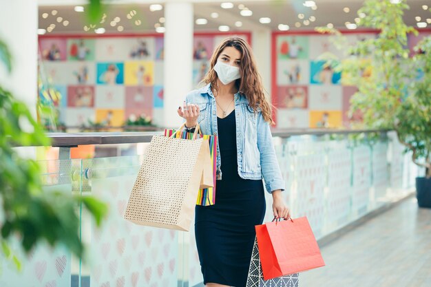 Mulher jovem feliz na máscara médica, caminhando no shopping com muitos sacos de compras nas mãos.