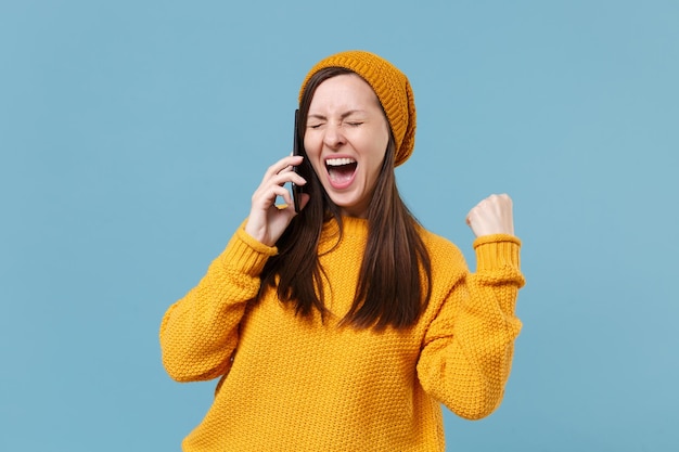 Mulher jovem feliz no chapéu de suéter amarelo posando isolado no retrato de estúdio de fundo azul. Conceito de estilo de vida de emoções sinceras de pessoas. Mock up espaço de cópia. Falando no celular fazendo gesto de vencedor.