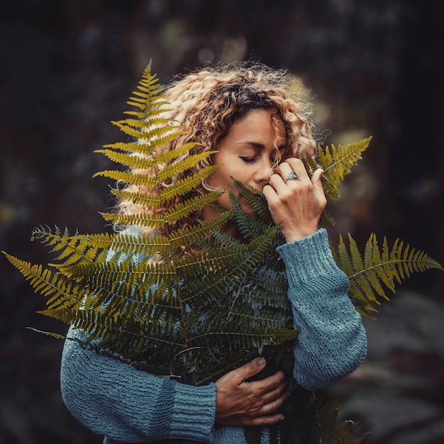 Mulher jovem feliz no suéter azul segurando samambaia fresca frágil deixa planta na floresta ou parque. Mulher jovem satisfeita segurando as folhas da planta. Mulher segurando uma folha de samambaia nas mãos, cobrindo parte do rosto