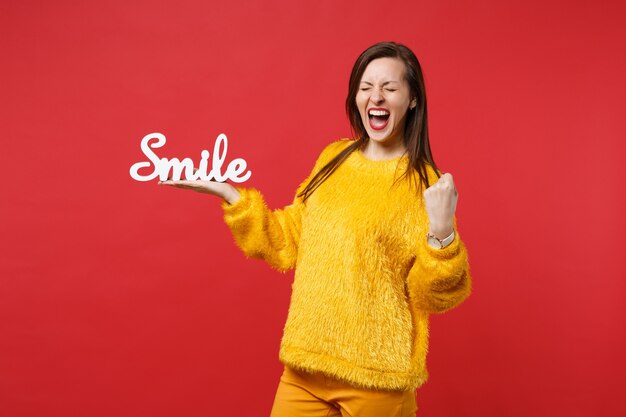 Mulher jovem feliz no suéter de pele com os olhos fechados, gritando o punho cerrado como o vencedor segurar o sorriso de letras de palavras de madeira isolado no fundo vermelho. conceito de estilo de vida de emoções de pessoas. simule o espaço da cópia.