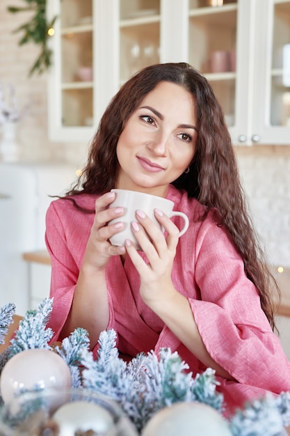 Mulher jovem feliz num vestido rosa com copo na cozinha decorada de Natal. morena linda com uma xícara de chocolate na cozinha com uma decoração de Natal. Luz branca cozinha interior para o Natal. Mulher em casa