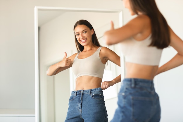 Mulher jovem feliz posando de jeans e aprovando novo tamanho gesticulando polegares para cima e sorrindo em pé perto do espelho