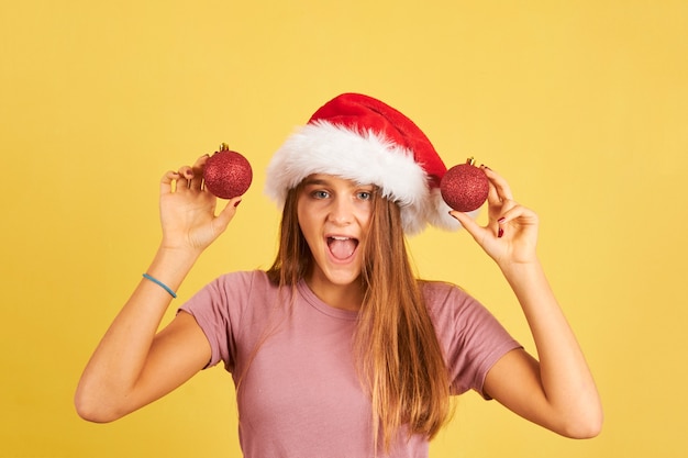 Mulher jovem feliz segurando bolas vermelhas de Natal e chapéu de Papai Noel em um fundo amarelo