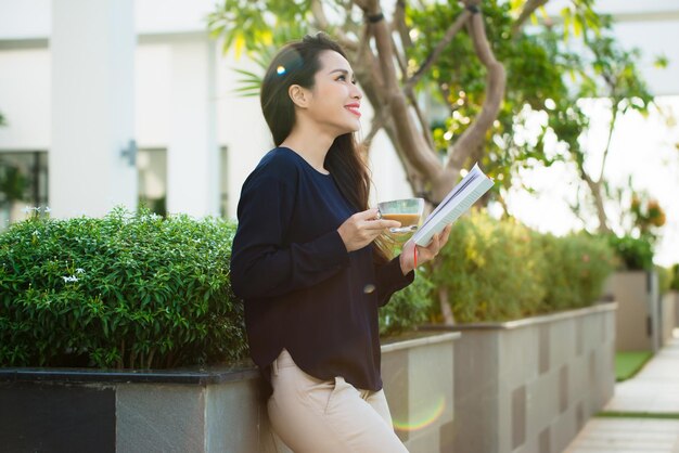 Mulher jovem feliz segurando o livro de literatura, analisando o romance durante o tempo de lazer no terraço do café do campus em dia ensolarado.