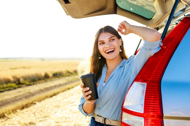 Mulher jovem feliz segurando uma xícara de café e sentada no porta-malas aberto de seu carro em um campo de trigo ao pôr do sol