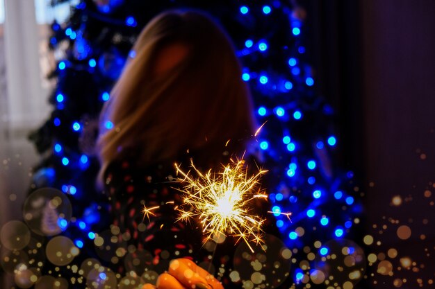 Mulher jovem feliz sentada com taça de champanhe e luz de Bengala ao lado da árvore de Natal. Celebração da festa de ano novo. Férias de inverno do Natal.