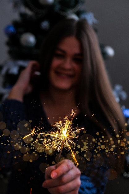 Foto mulher jovem feliz sentada com um copo de champanhe e luz de bengala ao lado da árvore de natal nova