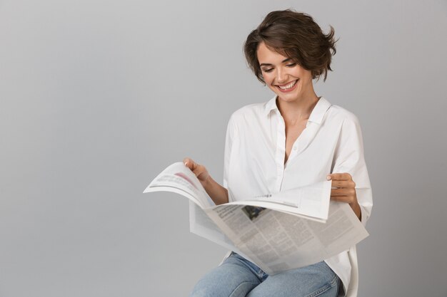 Foto mulher jovem feliz sentada em um banquinho, isolada na parede cinza, segurando um jornal