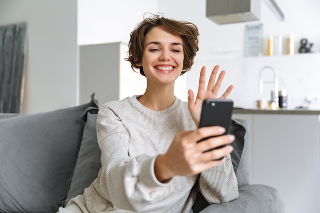 Mulher jovem feliz sentada em um sofá em casa, usando o celular, fazendo uma videochamada