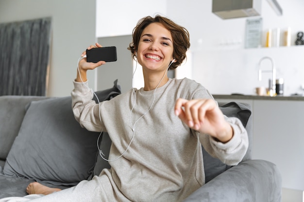 Mulher jovem feliz sentada em um sofá em casa, usando o celular, ouvindo música com fones de ouvido