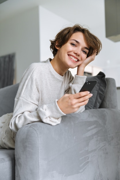 Mulher jovem feliz sentada em um sofá em casa, usando o celular