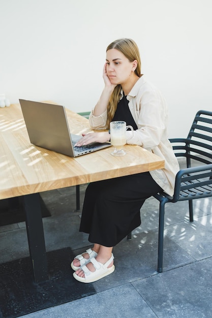 Mulher jovem feliz sentada em uma mesa de café ao ar livre e falando ao telefone com uma xícara de café, mulher sorridente desfrutando de teletrabalho em um café ou estudando on-line em um laptop