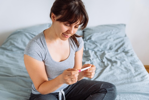 Mulher jovem feliz sentada na cama olhando para o teste de gravidez positivo