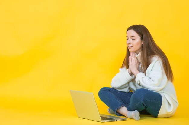 Mulher jovem feliz sentada no chão com as pernas cruzadas e usando laptop em amarelo