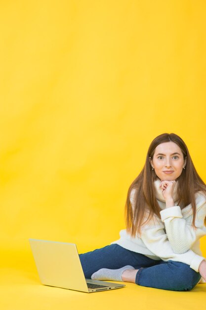 Mulher jovem feliz sentada no chão com as pernas cruzadas e usando laptop em amarelo