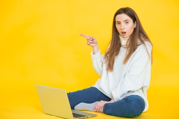 Mulher jovem feliz sentada no chão com as pernas cruzadas e usando laptop em amarelo