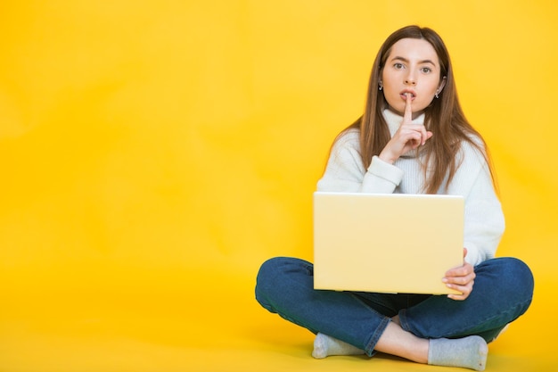 Mulher jovem feliz sentada no chão com as pernas cruzadas e usando laptop em amarelo