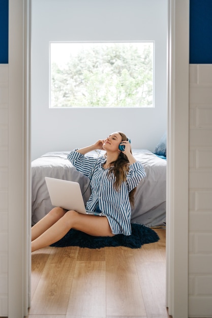 mulher jovem feliz sentada no quarto navegando no notebook enquanto ouve música