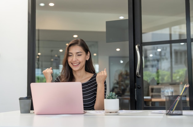 Mulher jovem feliz sorrindo e levantando as mãos enquanto olha para o laptop no escritório