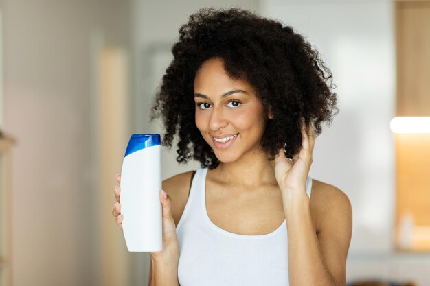 Mulher jovem feliz tocando o cabelo e segurando a garrafa de xampu.