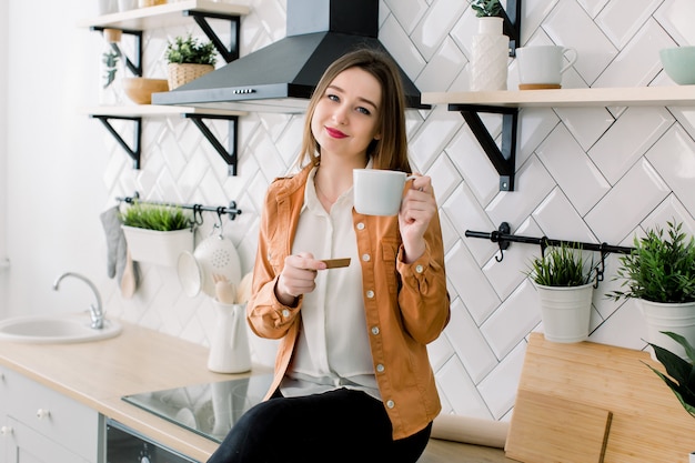 Mulher jovem feliz tomando café enquanto está sentado na cozinha em casa pela manhã