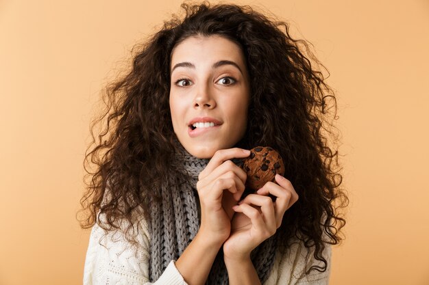 Foto mulher jovem feliz usando cachecol de inverno em pé isolado sobre uma parede bege, comendo biscoito de chocolate