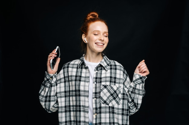Mulher jovem feliz usando fones de ouvido sem fio segurando o telefone e dançando ouvindo música online