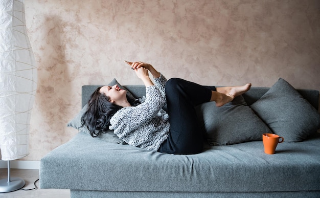 Foto mulher jovem feliz usando telefone móvel enquanto deitada no sofá em casa