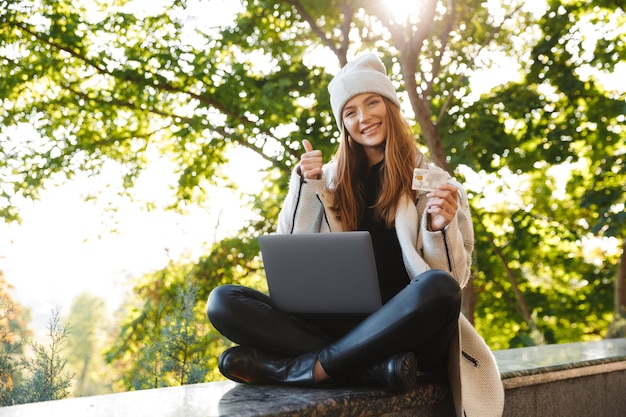 Mulher jovem feliz vestida com um casaco e chapéu outono sentada ao ar livre, usando um computador laptop, mostrando um cartão de crédito de plástico
