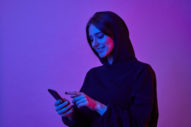 Mulher jovem feliz vestindo um capuz e usando o smartphone em luzes de néon. Menina navegando na internet no telefone em uma luz de néon rosa.