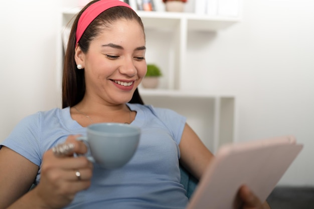 Mulher jovem feliz visualizando suas redes sociais em um tablet digital bebendo uma xícara de café