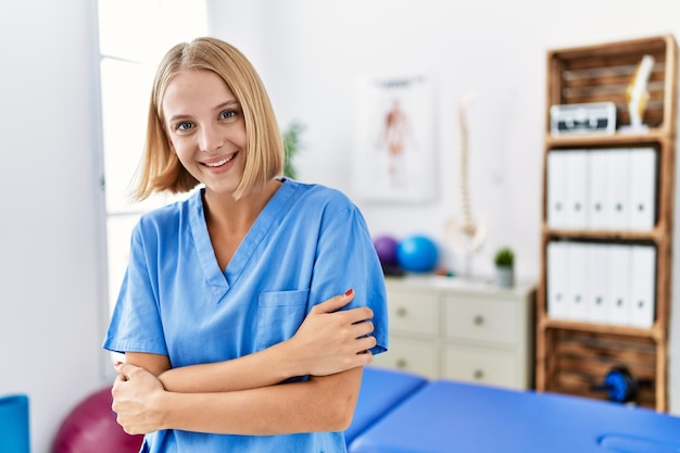 Mulher jovem fisioterapeuta caucasiana trabalhando na clínica de recuperação de dor rosto feliz sorrindo com os braços cruzados olhando para a câmera pessoa positiva
