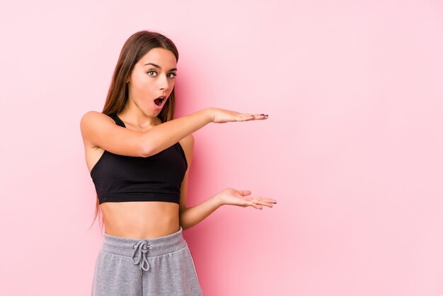 Mulher jovem fitness caucasiano posando em uma parede rosa chocada e espantada, segurando um espaço de cópia entre as mãos.