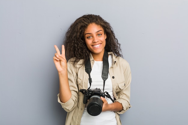 Mulher jovem fotógrafo americano africano segurando uma câmera mostrando o número dois com os dedos.